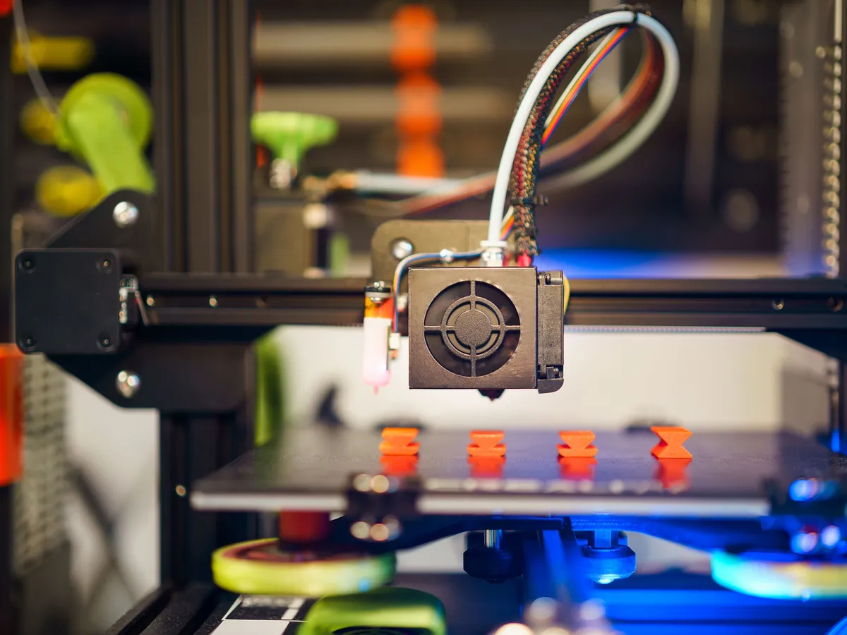 Close-up of a 3D printer extruder over a print bed with small orange printed parts