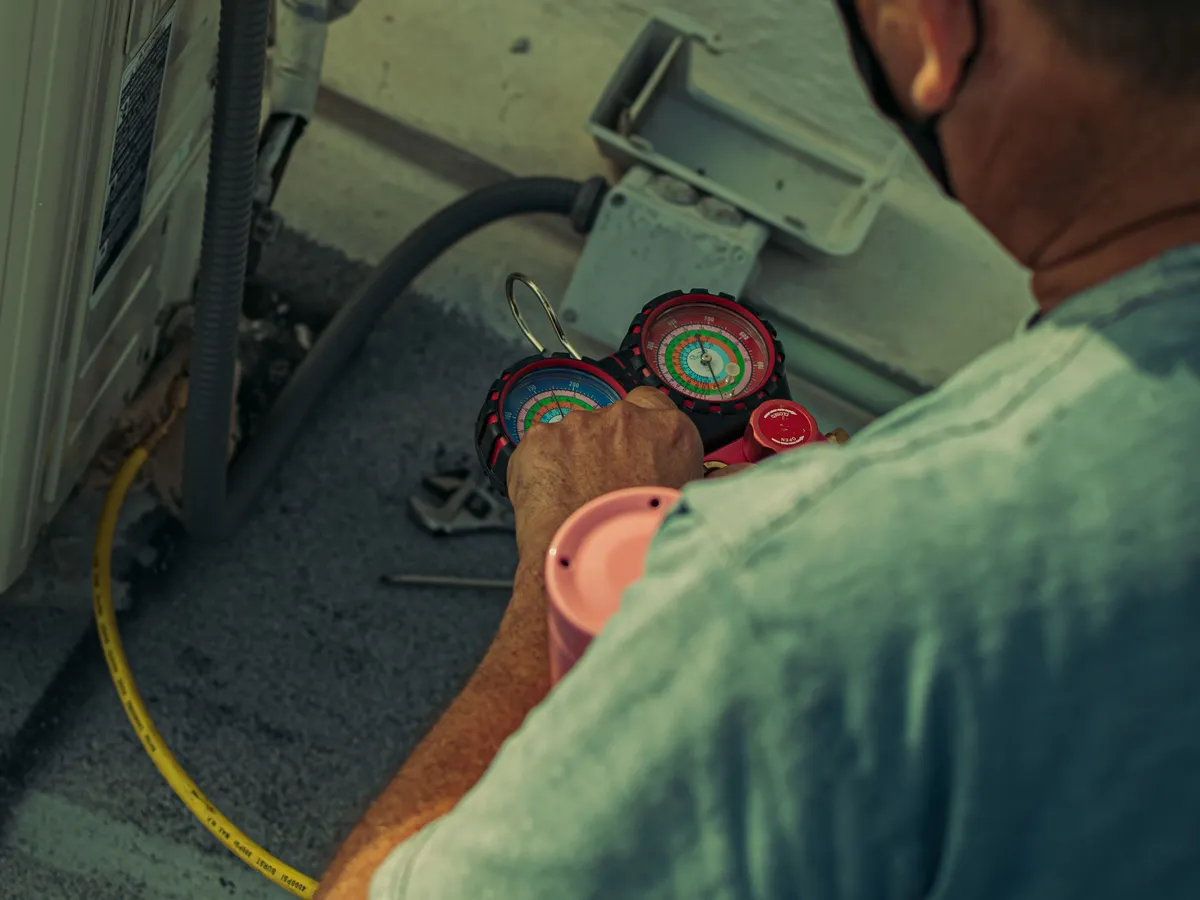 HVAC technician in a face mask reading refrigerant manifold gauges beside an outdoor AC unit