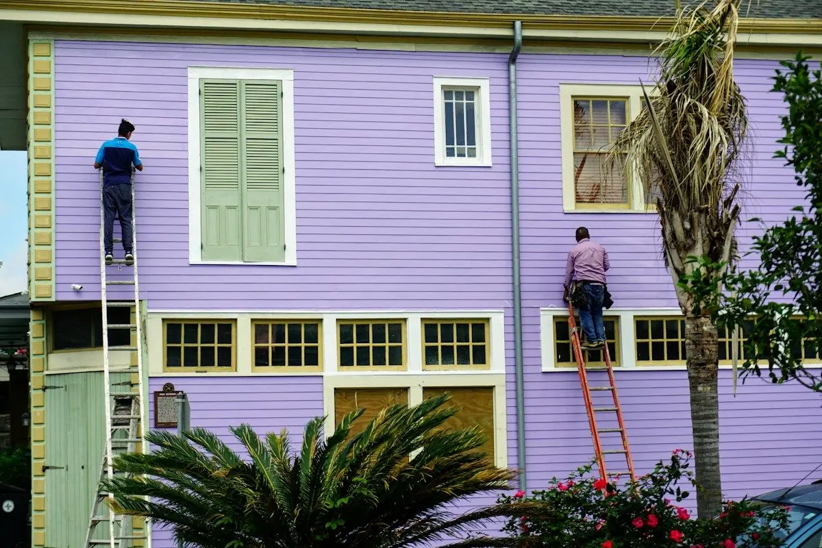 Two workers on ladders painting and repairing the siding of a two-story house