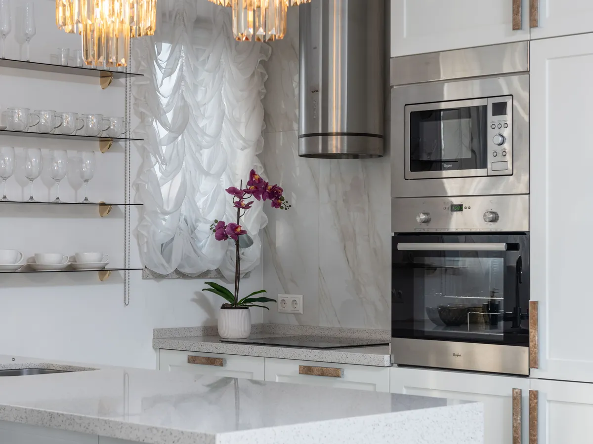 Built-in stainless steel microwave above an electric oven in white cabinetry beside a kitchen window with a sheer curtain