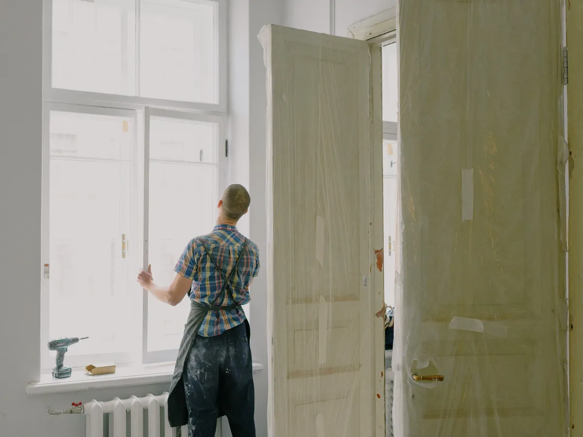 Installer in a plaid shirt and apron standing at a tall white window frame beside a drill and a protected door