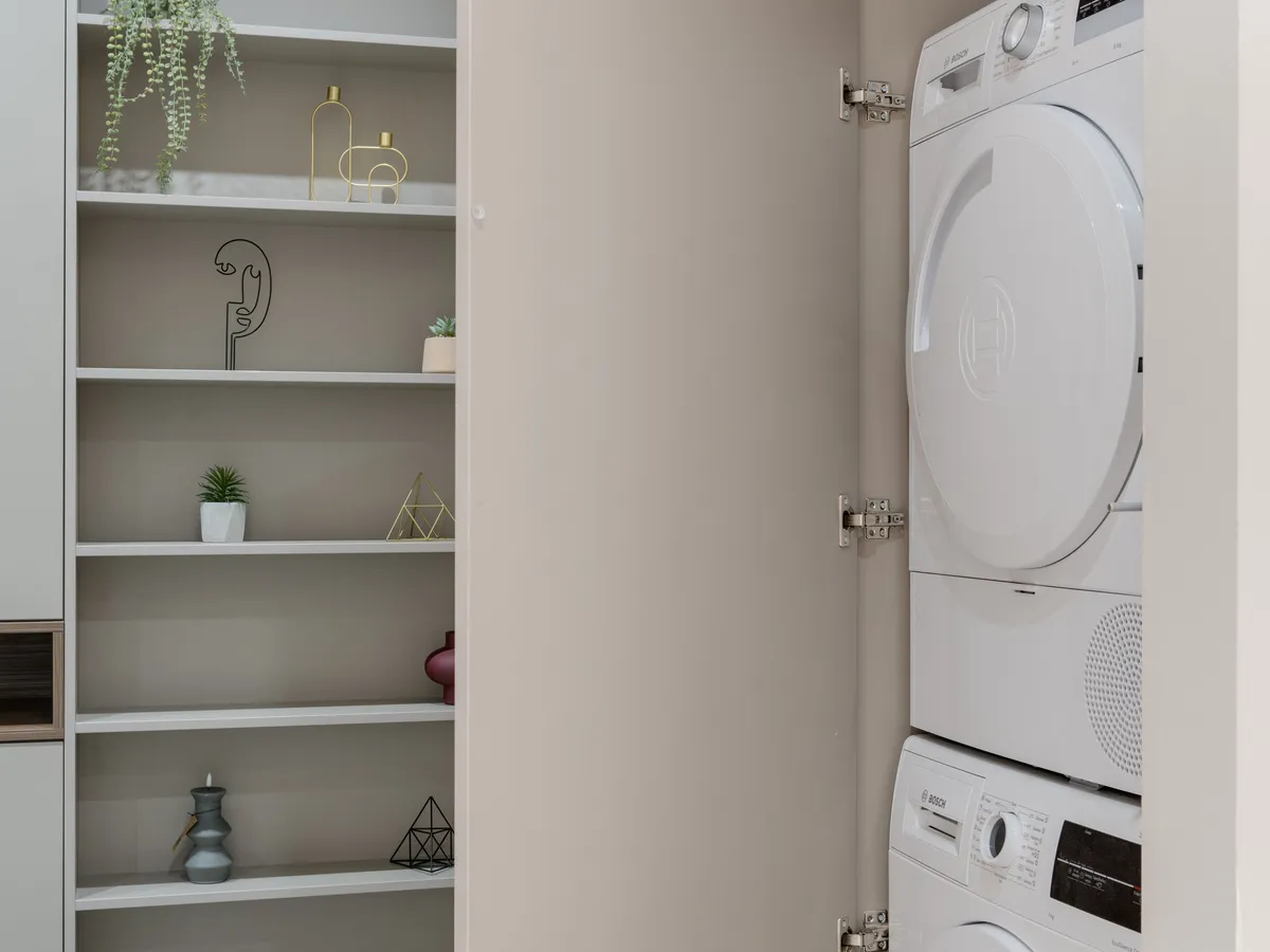 White front-load dryer stacked on top of a matching washer inside a cabinet alcove, next to shelves