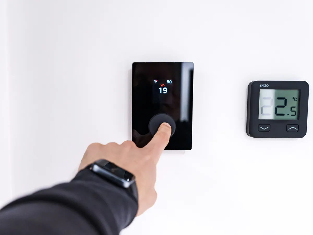 Hand pressing the dial of a wall-mounted digital thermostat next to a second small display