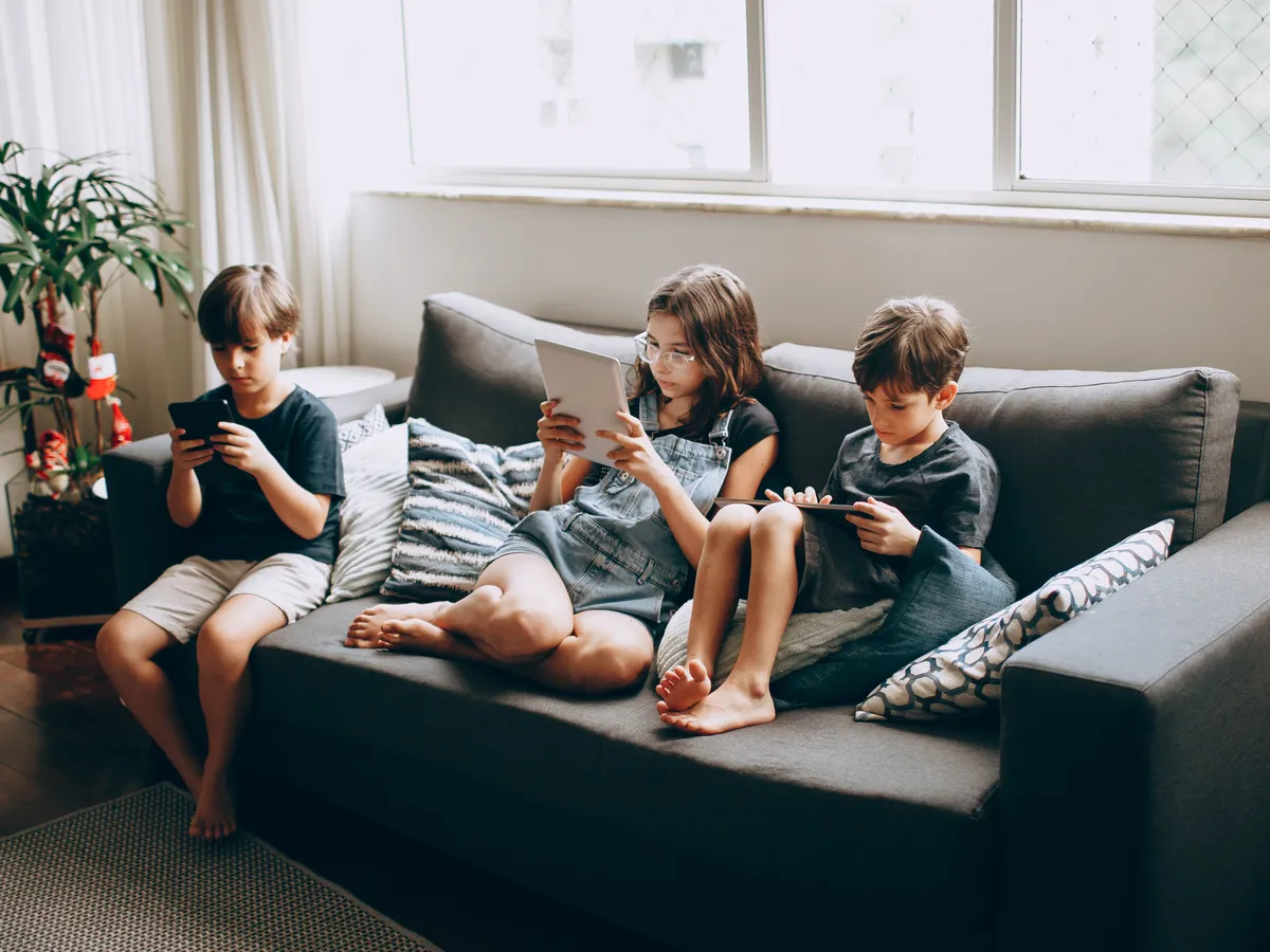 Three children sitting on a sofa using tablets and phones near a window
