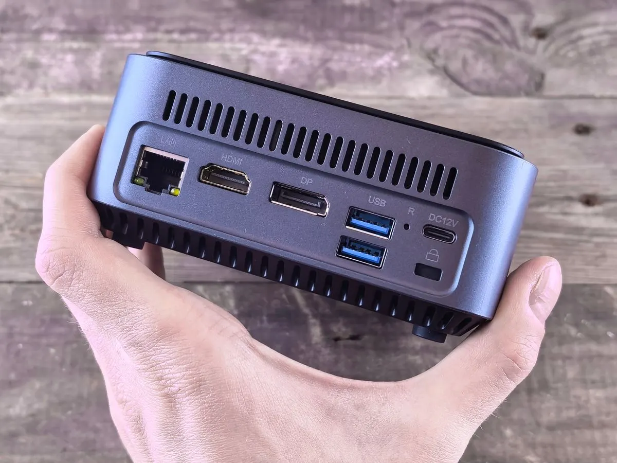 Hand holding a compact gray mini PC showing the LAN, HDMI, DisplayPort, USB and power ports on its back panel