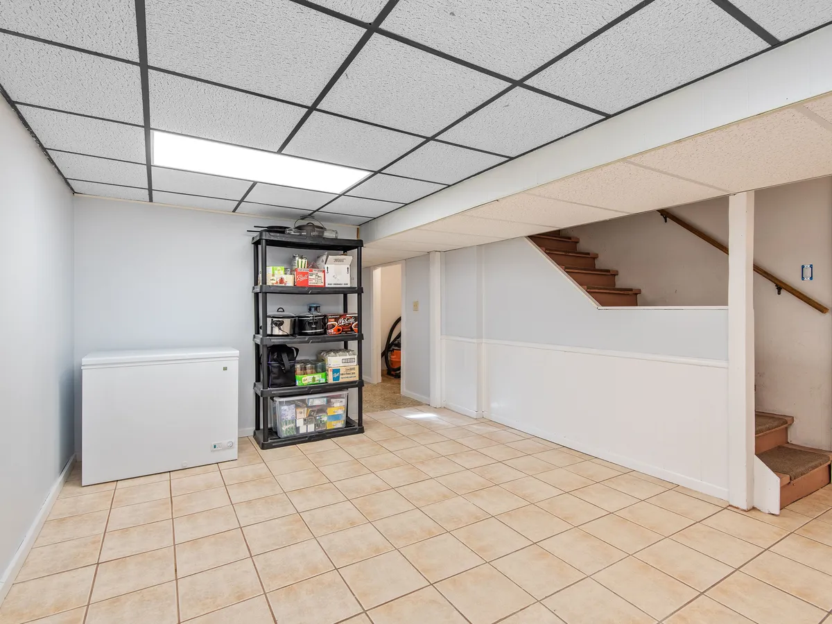 Finished basement room with a drop ceiling, tile floor, a chest freezer, shelving and a staircase