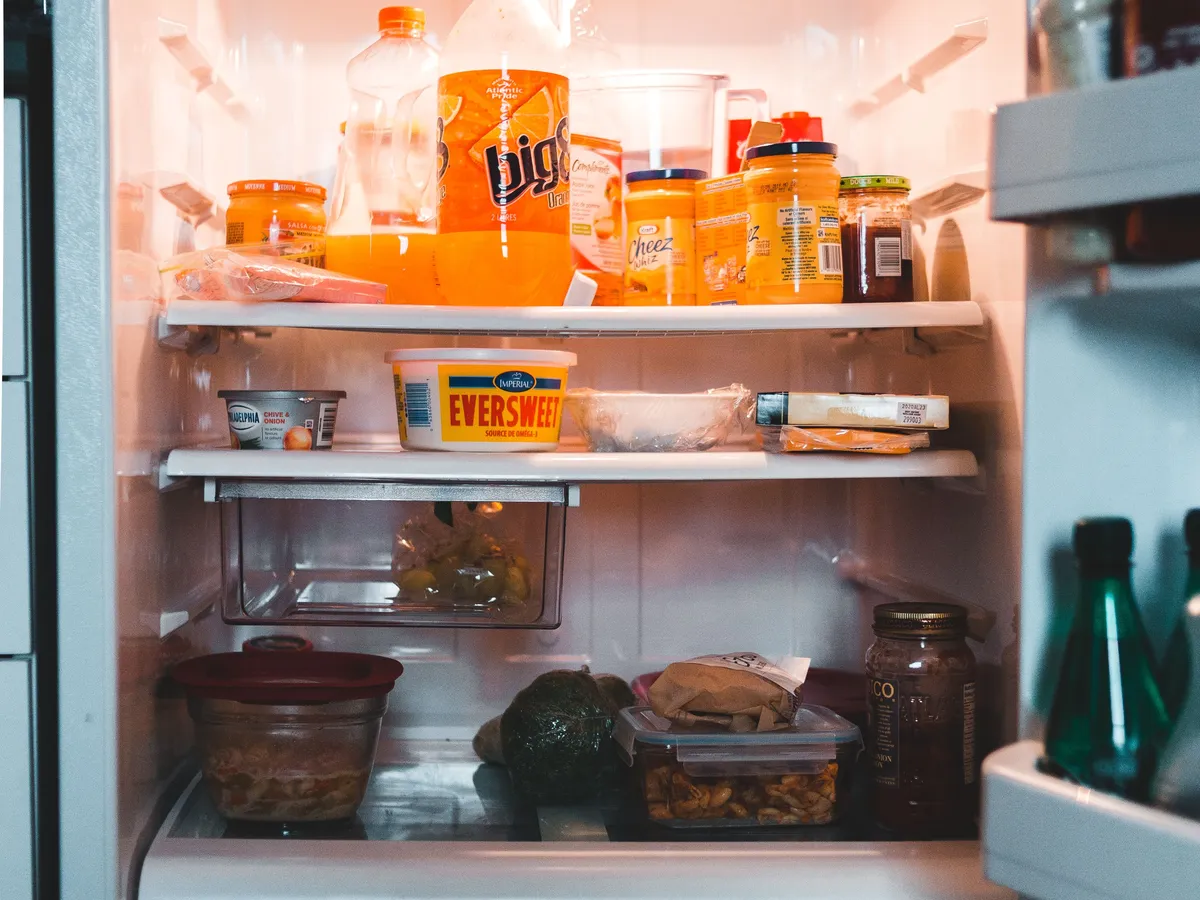 Open refrigerator interior lit from inside, with shelves of orange juice bottles, spreads, containers and an avocado