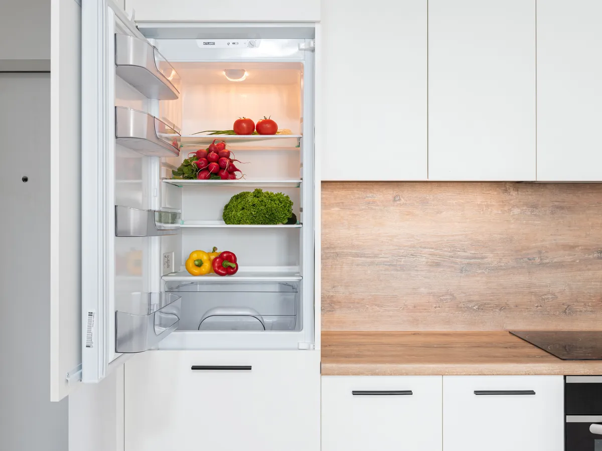 Open white refrigerator with a door that swings out, holding tomatoes, radishes, lettuce and bell peppers in a kitchen with white cabinets
