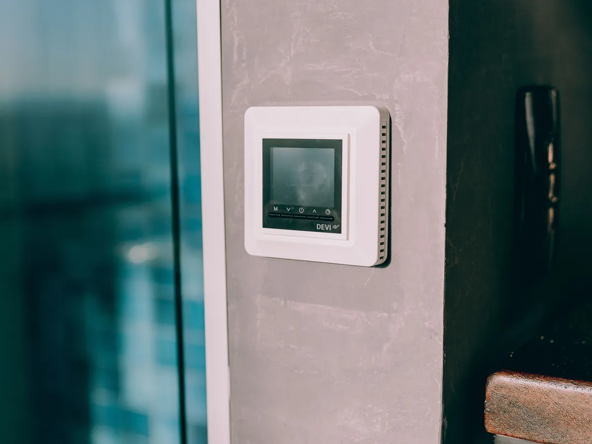 White digital wall thermostat mounted on a gray concrete wall beside a window