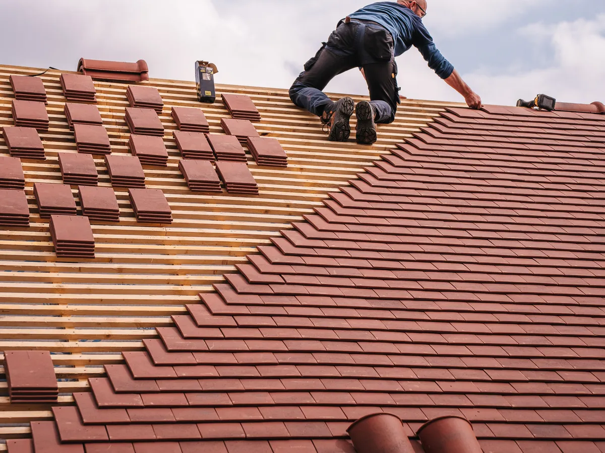 Roofer kneeling on a pitched roof laying red clay tiles over wooden battens