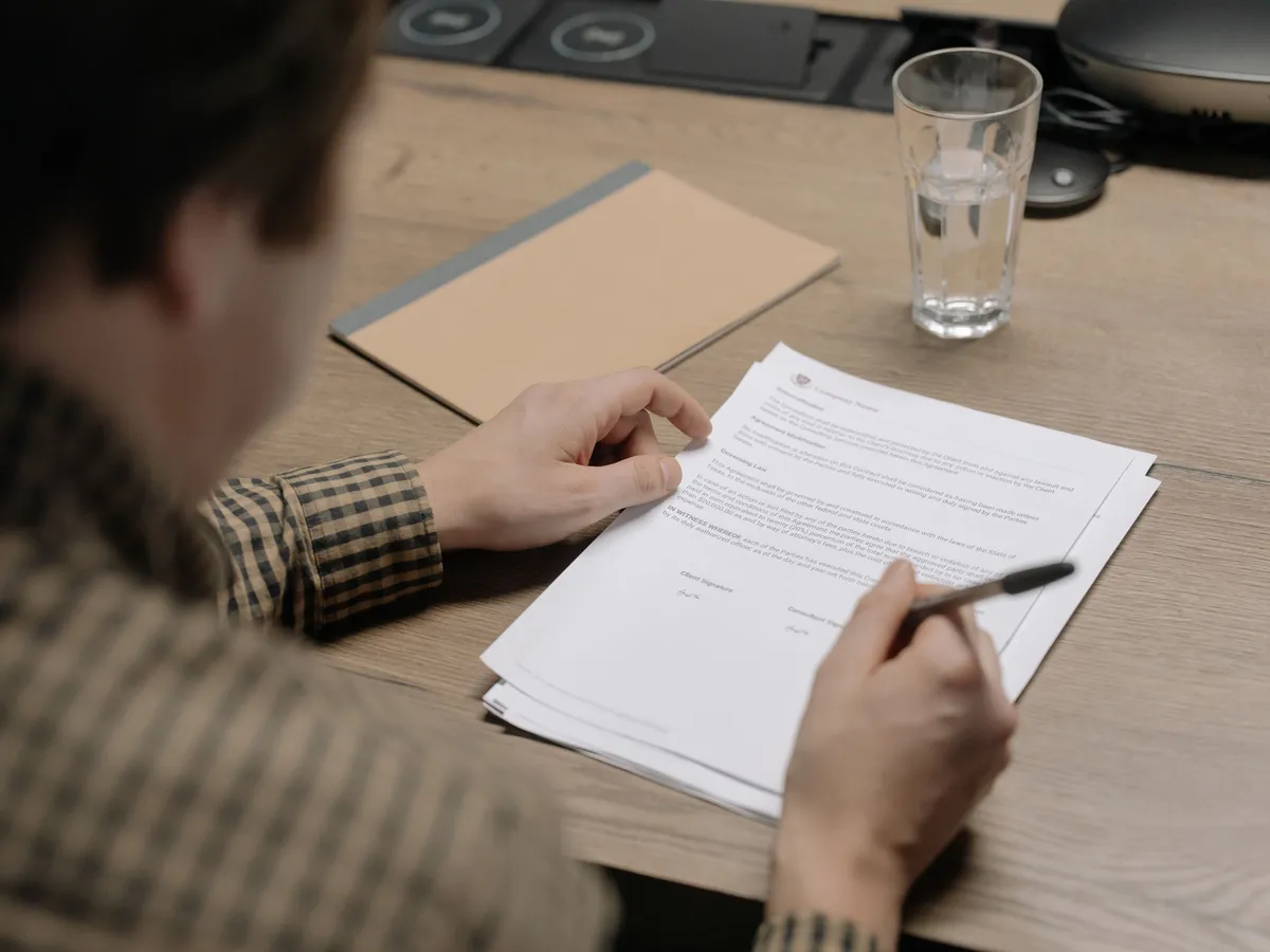 Person in a checked shirt holding a pen over a printed contract on a wooden table with a notebook and glass of water