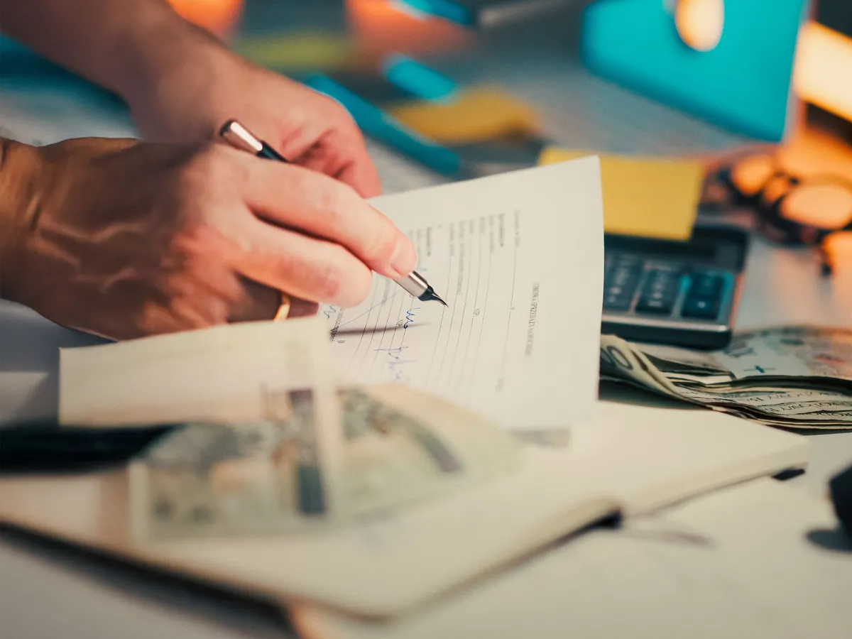 Hand holding a pen over a signed form on a desk with a calculator, notebook and banknotes nearby