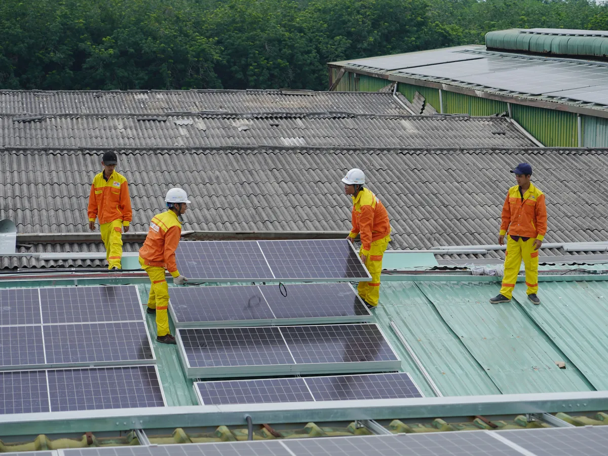 Four workers in orange coveralls carrying solar panels across a corrugated roof next to rows already installed