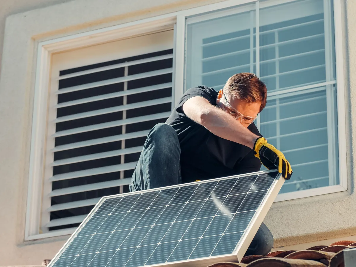 Installer in safety glasses and yellow work gloves lifting a solar panel up onto a tile roof beside a window