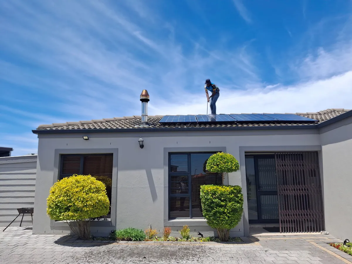 Worker standing on the roof of a single-story house in front of a row of solar panels under a bright blue sky