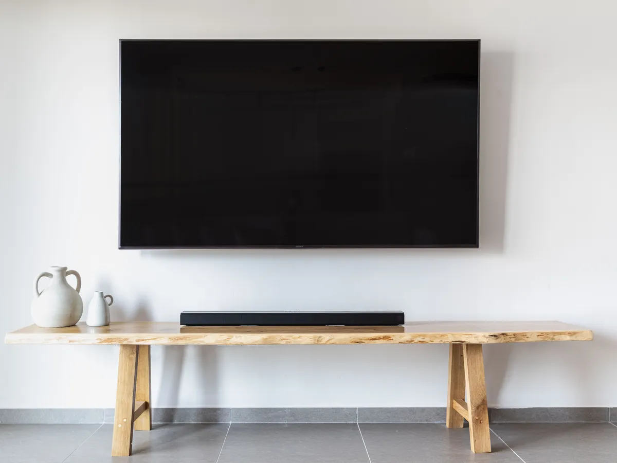 Large flat-screen TV mounted on a white wall above a wooden bench and soundbar