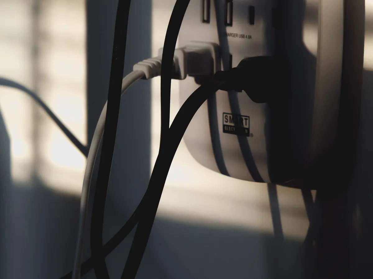 Close-up of black and white cables plugged into a wall power strip, backlit by sunlight through blinds