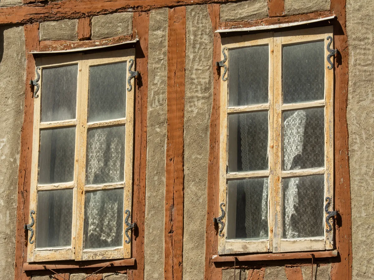 Two old wooden-framed windows with six panes, iron hinges, and lace curtains set in a half-timbered wall