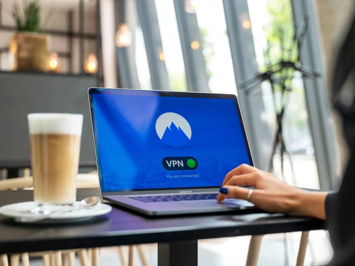 Laptop on a cafe table showing a VPN app with a green connected indicator next to a glass of iced latte