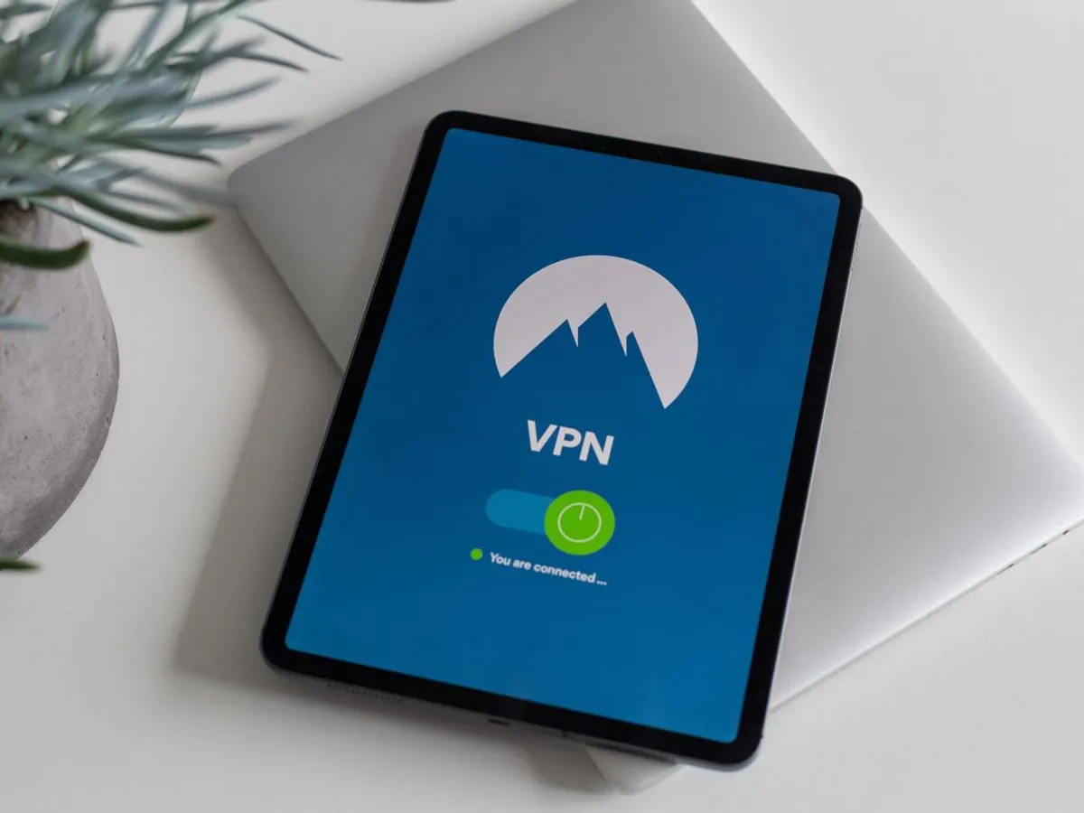 Tablet on a desk showing a VPN app with a green connected toggle, resting on a closed laptop beside a potted plant